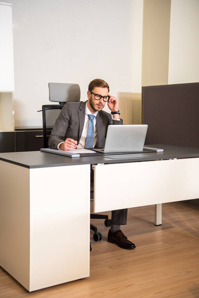 Joven hombre de negocios usando el ordenador portátil mientras está sentado en la mesa en la oficina
 - Foto, Imagen