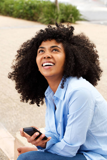 Εσωτερικη πορτρέτο του χαρούμενος νεαρός αφρικανική αμερικανική γυναίκα που κάθεται σε εξωτερικούς χώρους με κινητό τηλέφωνο - Φωτογραφία, εικόνα