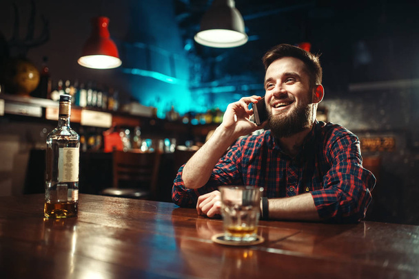 jonge man met fles whisky bij bar teller en praten over mobiele, alcoholisme, dronkenschap concept - Foto, afbeelding