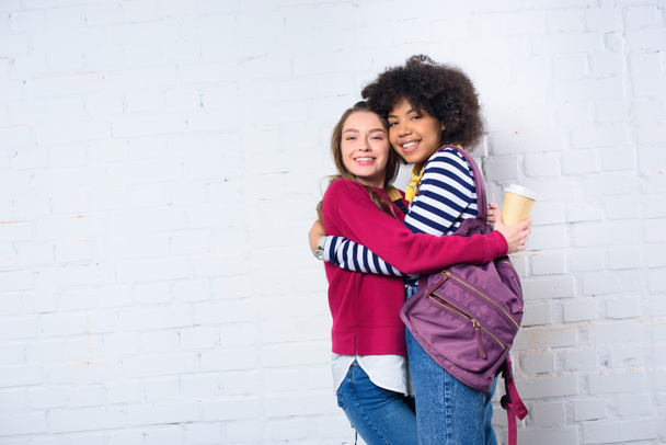 portrait of cheerful multicultural students hugging each other against white brick wall - Fotografie, Obrázek