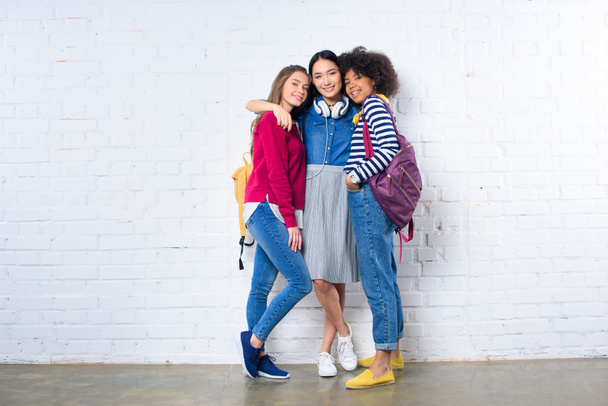 asian student hugging smiling multiethnic friends against white brick wall - Foto, afbeelding