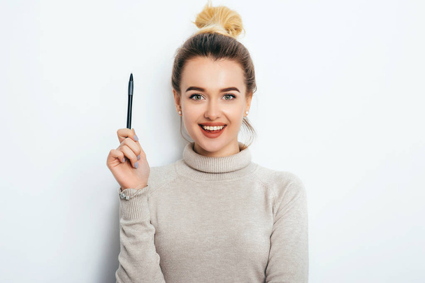 Portrait of cheerful woman with appealing smile, having hair bun in sweater isolated on white background holding pen and have a great idea. Beautiful female showing emotions. People Beauty Fashion - Photo, Image