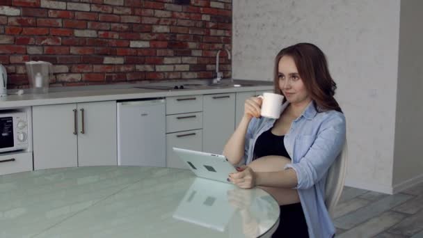 Счастливая беременная женщина сидит за стеклянным кухонным столом, пьет кофе и пользуется планшетным компьютером. делает онлайн-покупки для новорожденных
 - Кадры, видео