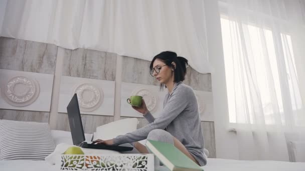 Cheerful girl in eyeglasses typing on laptop computer for sharing social media sitting in bed and drinking coffee at home in the morning - Metraje, vídeo
