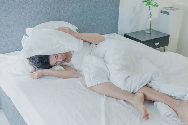 Young caucasian woman with black hair sleeps funny in bed, covering her head with pillow. Noisy neighbors interfere, lack of sleep, too lazy to get out of bed. - Foto, imagen