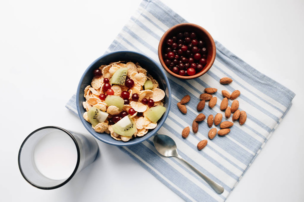 glass of milk, delicious crispy cornflakes with kiwi pieces and cranberries in bowl, almonds on dish cloth, closeup, healthy breakfast  - Photo, Image