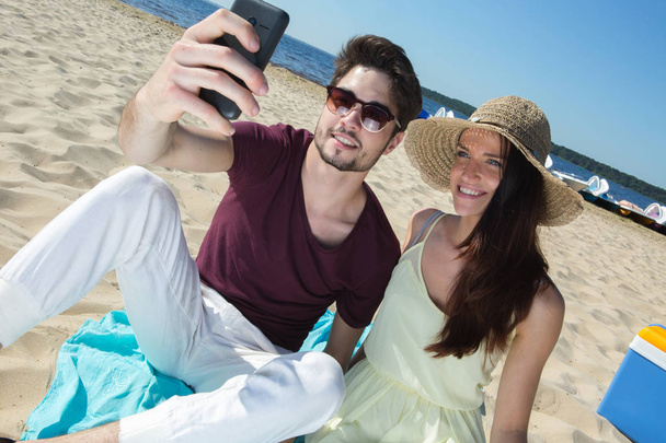 gorgeous young couple sitting at the beach and doing selfie - Foto, afbeelding