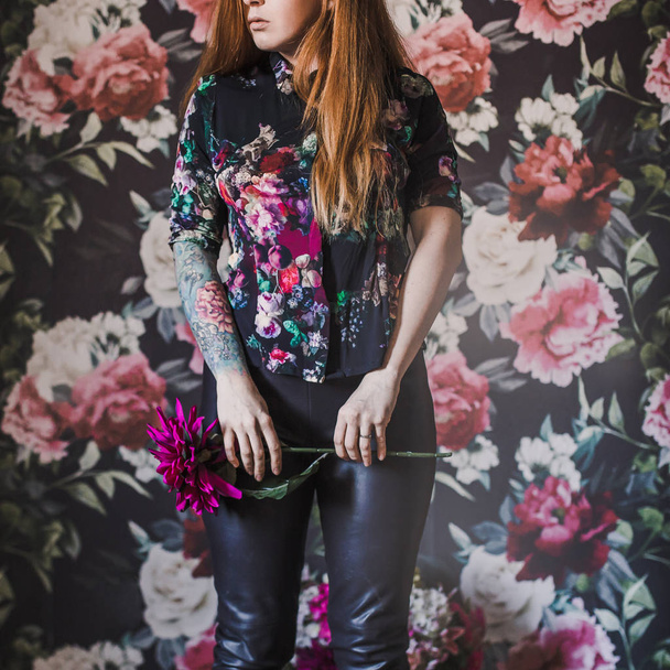 young woman with long red hair in leather pants holding pink flower on colored wall with flowers - Photo, Image