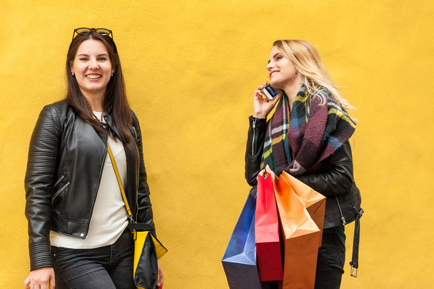 2 つのかわいい女の子ブルネットとブロンド金曜日黒地ショッピングの日があります。それらの 1 つ、携帯電話に話している他の笑顔。プレーンの黄色の壁の前で - 写真・画像