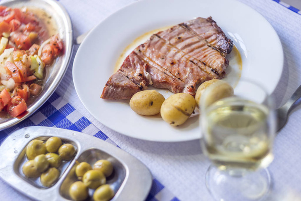 Tuna steak accompanied with potatoes, olives, tomato salad and w - Photo, Image