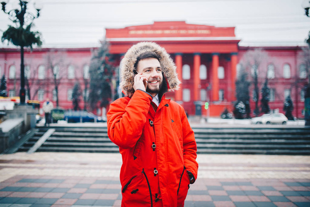 Sakal ve kırmızı ceket başlıklı bir öğrenci genç adam cep telefonu kullanır, üniversite veya kolej bina kırmızı zemin üzerine gülümseme ile telefonda konuşurken yakınındaki baş, elinde tutan - Fotoğraf, Görsel