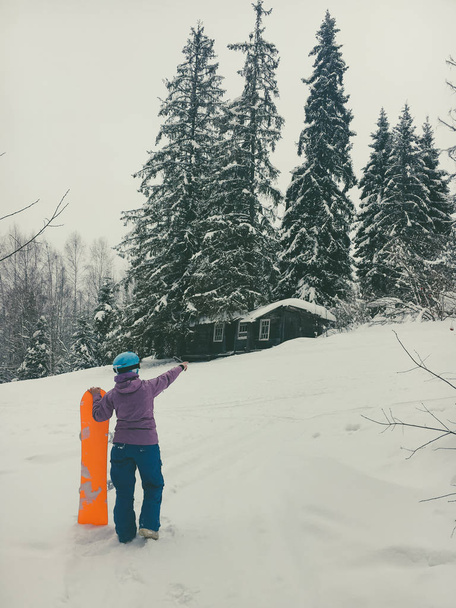 сноубордистка в синем шлеме стоит спиной держа доску рядом с ярко-оранжевой доской и показывает направление вперед в лесу и смотрит на заброшенный старый деревянный домик на поляне
 - Фото, изображение