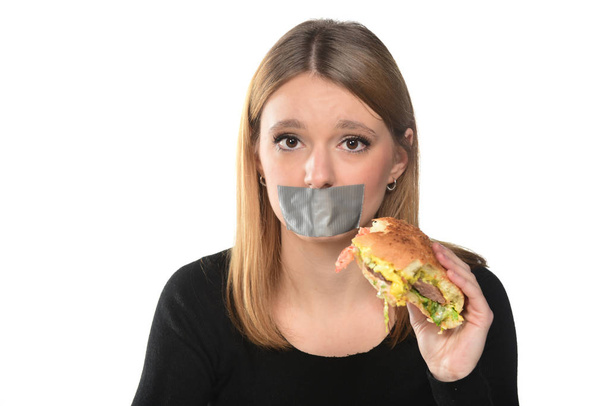 portrait of a young girl with hamburger in hand and tape over her mouth on the white background - Photo, image