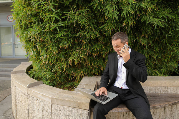Manageri puhuu älypuhelimella ja työskentelee kannettavan tietokoneen kanssa ulkona
 - Valokuva, kuva