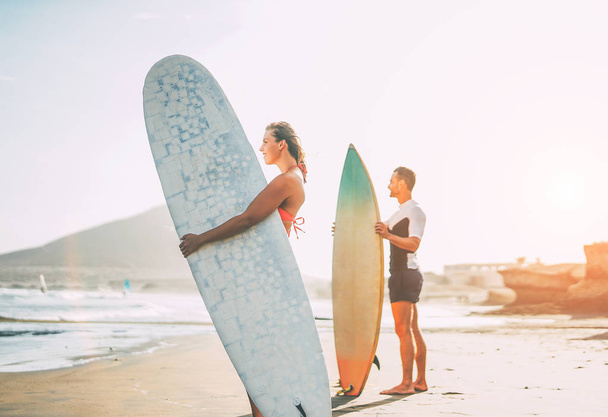 Young couple of surfers standing on the beach with surfboards preparing to surf on high waves during a magnificent sunset - People, lifestyle, sport concept - Focus on woman - Photo, Image