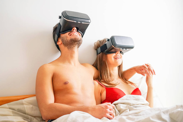 glückliches Liebespaar mit Virtual-Reality-Brille im Bett unter weißen Laken - verliebte junge Romantiker mit vr-Simulationsvision im Schlafzimmer - neues technologisches Lifestyle-Konzept - Foto, Bild