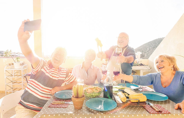 Happy seniors taking a selfie while making a barbecue on the rooftop at sunset - Retired people having fun eating and drinking red wine laughing together - Cheerful elderly happiness concept - Photo, Image