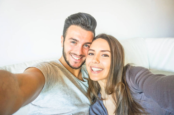 Happy young couple taking a selfie with mobile smart phone camera in the living room embracing on sofa at home - Friends making self portrait on the couch - People, relationship, lifestyle concept - Φωτογραφία, εικόνα