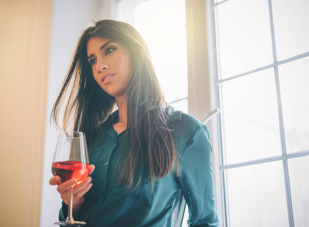 Gelukkig jonge vrouw in haar appartement drinken van een glas rode wijn vervolgens het venster met een schattige glimlach - vrouw genieten van haar tijd thuis met een cocktail - plezier, entertainment en lifestyle concept - Foto, afbeelding