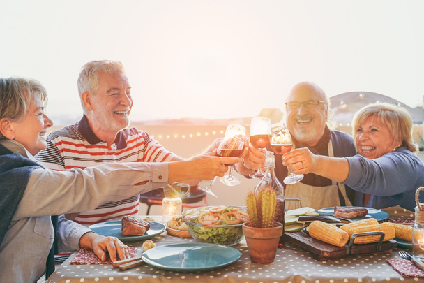 Szczęśliwy starszy przyjaciele zabawy doping z czerwonego wina na grillu w taras odkryty - dojrzałe osoby czynią kolacji opiekania okulary i śmiejąc się razem - przyjaźń i koncepcja starszych styl życia - Zdjęcie, obraz