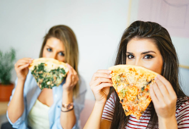 Nuoret kauniit naiset syövät makupaloja italialaista pizzaa kotona Iloiset kauniit sisaret peittävät kasvonsa pikaruoalla asunnossa Ihmisten käsite, elämäntapa, ateria Keskity naisen silmään
 - Valokuva, kuva
