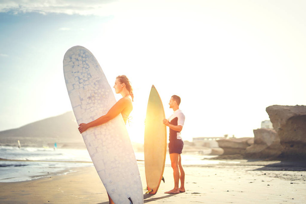 Young couple of surfers standing on the beach with surfboards preparing to surf on high waves during a magnificent sunset behind the mountain - People, lifestyle, sport concept - Focus on woman - Photo, Image