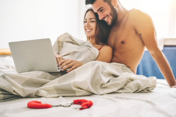 Happy νεαρό ζευγάρι ξαπλωμένο σε ένα κρεβάτι με υπολογιστή - όμορφη παντρεμένο ζευγάρι βλέποντας ρόλο παιχνίδια σεξ βίντεο στον φορητό υπολογιστή που γελά μαζί - ανθρώπων, σεξουαλική, τεχνολογία έννοια - μαλακή εστίαση σε φύλλα - Φωτογραφία, εικόνα