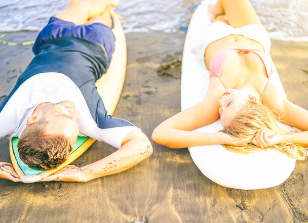 Ευτυχισμένο ζευγάρι των surfers ξαπλωμένη για ιστιοσανίδες στην παραλία το ηλιοβασίλεμα για χαλάρωση μαζί - υγεία φίλους γέλιο κοιτάζοντας κάθε τους άλλους και να απολαύσετε την ημέρα μετά από τις αθλητικές δραστηριότητες - Φωτογραφία, εικόνα