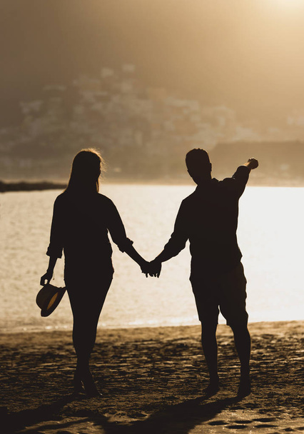 Вид сзади красивой молодой пары подростков, держащихся за руки на пляже во время заката - Силуэт двух влюбленных, имеющих романтический момент на пляже - Концепция любви и отдыха - Фото, изображение