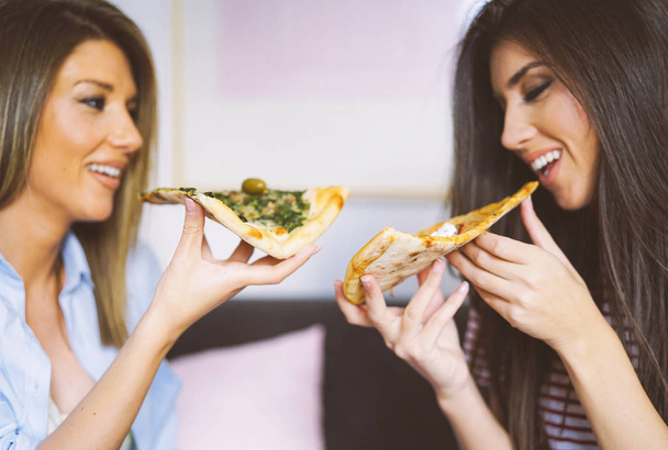 Nuoret kauniit naiset syövät viipaleita maukasta italialaista pizzaa kotona Iloiset kauniit naiset nauttivat nopean aterian yhdessä asunnossaan Ihmisten käsite, elämäntapa, ruoka Keskity pizzaan
 - Valokuva, kuva