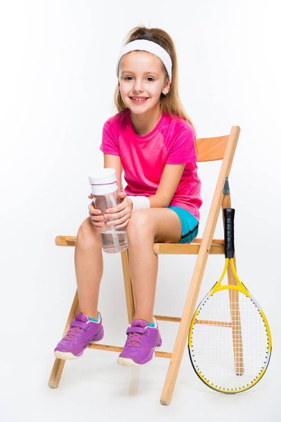 Niedliche kleine Tennisspielerin sitzt auf einem Holzstuhl und hält eine Flasche Wasser in der Hand  - Foto, Bild