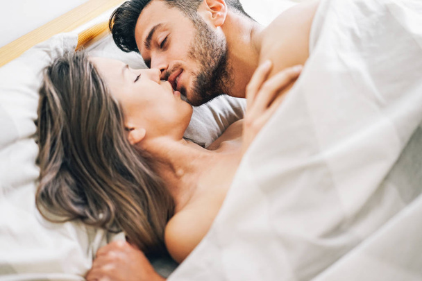 Jeune couple amoureux embrassant dans un lit sous couverture blanche - Amoureux passionnés ayant des moments romantiques et intimes sur le lit - Concept de sexe et de passion - Focus sur le visage masculin
 - Photo, image