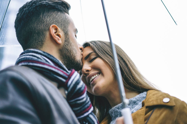 glückliches Paar küsst sich unter einem Regenschirm an einem regnerischen Tag - schöner Mann küsst seine Stirn Freundin unter dem Regen - Liebe, Menschen, Beziehungskonzept - Fokus auf die Frau - Foto, Bild