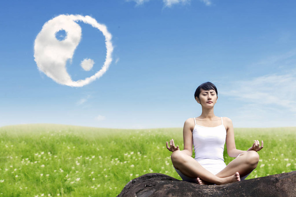 Asian woman meditating outdoors on the rock at meadow with yin yang clouds symbol in the sky - Фото, зображення