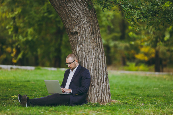 Mladý úspěšný podnikatel v bílé košili, klasický oblek, brýle. Muž sedí na trávě zem, práce na pc notebook v městském parku na zeleném trávníku venku v přírodě. Mobilní kancelář, obchodní koncept. - Fotografie, Obrázek