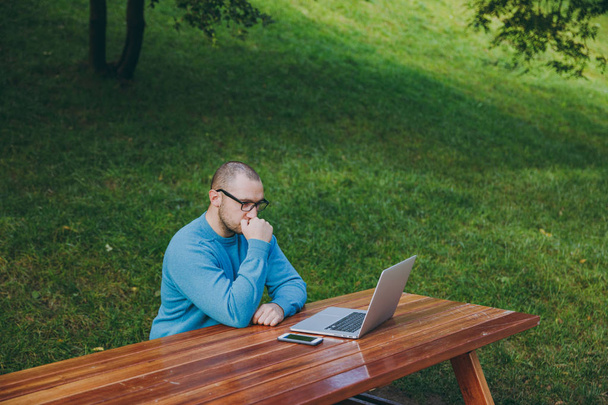 Young pensive successful smart man businessman or student in casual blue shirt, glasses sitting at table with mobile phone in city park using laptop, working on nature outdoors. Mobile Office concept. - Photo, Image