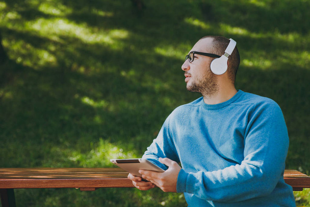 Young happy man, businessman or student in casual blue shirt glasses sitting at table with headphones, tablet pc in city park, listen music, rest outdoors on green nature. Lifestyle leisure concept. - Фото, зображення