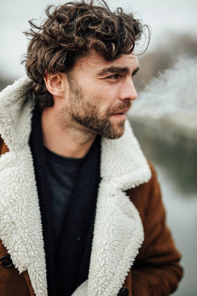 cool handsome man smoking cigarette outside in a aviator coat with masculine attitude waiting for some - Фото, зображення