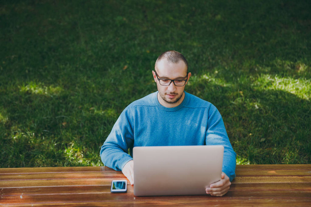 Young successful smart man businessman or student in casual blue shirt glasses sitting at table with mobile phone in city park using laptop working outdoors on green background. Mobile Office concept. - Fotó, kép