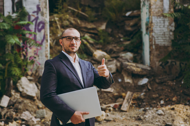 Successful businessman in white shirt, classic suit, glasses. Man show thumb up, stand with laptop pc computer phone near ruins, debris, stone building outdoors. Mobile Office, business, work concept. - Photo, Image