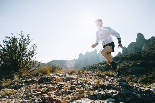 Professional trail runner, ultra distance athlete runs through rocky terrain high on mountain path or hiking trail, healthy lifestyle activity outdoors on sunny day. Active young man in sport wear - Foto, afbeelding