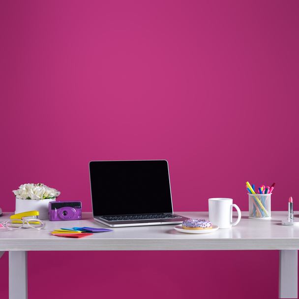 Laptop mit leerem Bildschirm, Donut mit Kaffee, Kamera, Kosmetik und Bürobedarf auf rosa - Foto, Bild