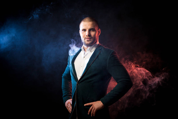 Young handsome man manager in white shirt and green jacket posing against red and blue smoke from a wipe on black isolated... - Foto, Imagen