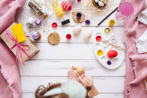Вид сверху на маленькую девочку, рисующую пасхальное яйцо с желтой краской и кистью
 - Фото, изображение