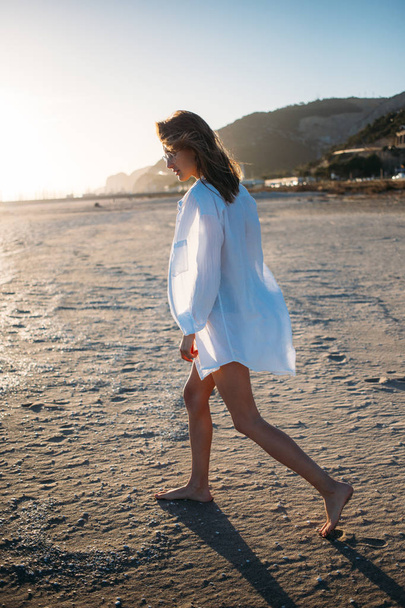 Beautiful and skinny young woman or girl models in one piece swimsuit and white linen shirt, walks in warm summer sunset light towards edge of water on empty beach, concept leisure and paradise - Foto, Imagen