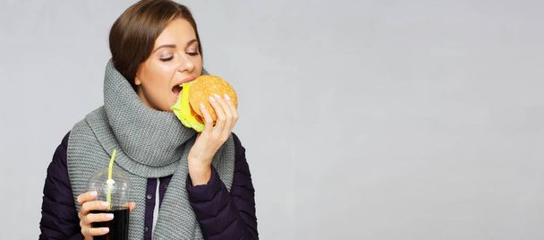 Woman wearing winter scarf biting burger and holding cola glass  - Foto, Imagen
