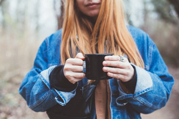 Cute young hipster with red hair and in blue denim oversize jacket holds black camping mug or cup with coffee during trip to forest or camp, nomad outfit lifestyle or travel inspiration - Photo, Image