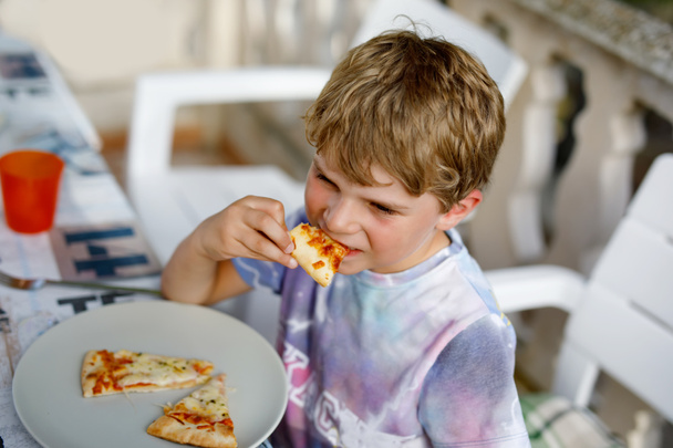 Cute healthy preschool kid boy eats fresh pizza sitting on terrace in summer, outdoors - Photo, Image
