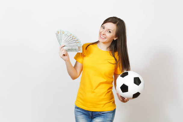 Joven mujer europea divertida, fanático del fútbol o jugador en uniforme amarillo sosteniendo montón de billetes de dinero, pelota de fútbol aislado sobre fondo blanco. Deporte, juego de fútbol, emoción concepto de estilo de vida
. - Foto, imagen