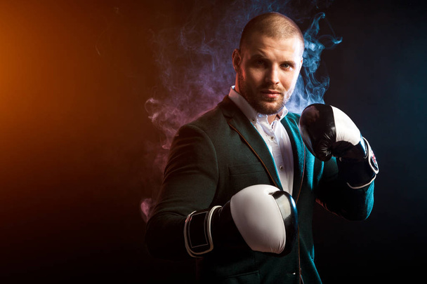 Young handsome businessman man in white shirt, green jacket, black and white boxing gloves smiling and ready to box against the red and blue smoke from the paper on a black isolated background - Foto, Imagem